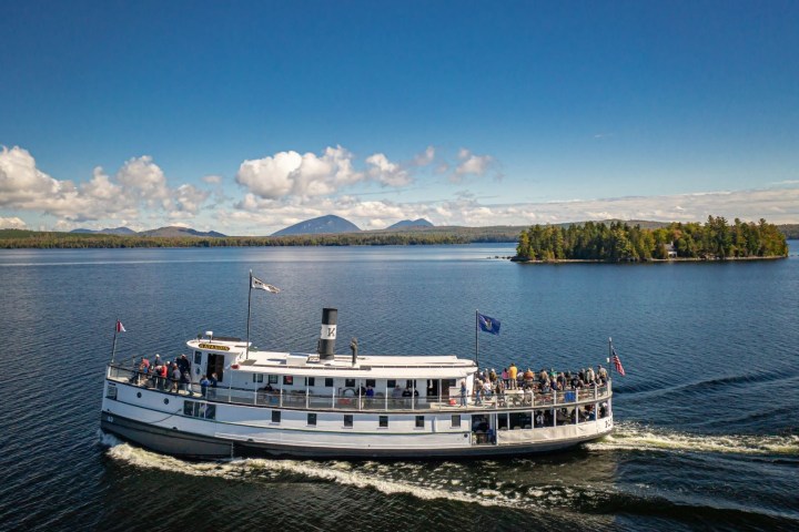 A white steamboat cruises on a lake with an island and mountains in the background.