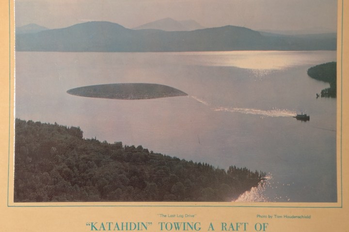 A boat towing a large log raft on a lake surrounded by forests and islands under a blue sky.