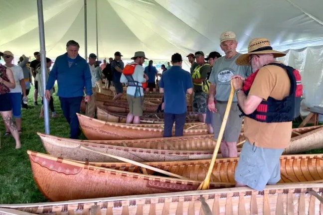 People gather under a tent inspecting wooden canoes displayed on grass.